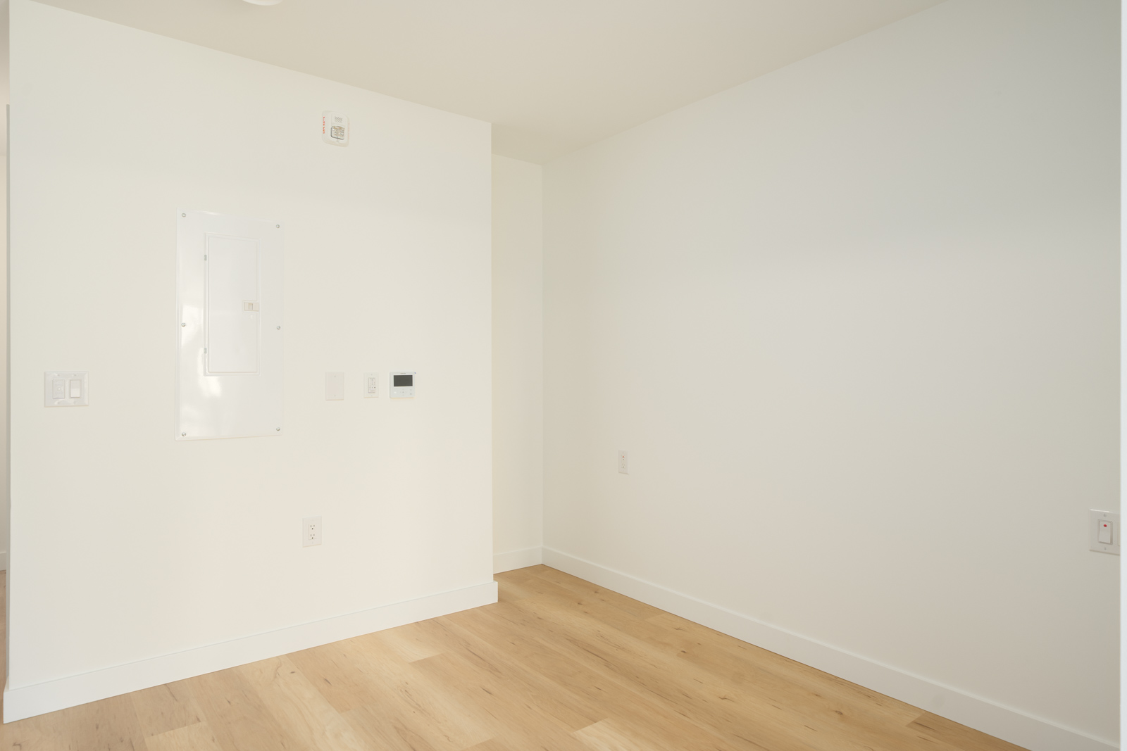 Empty 2nd-floor office corner in Port Coquitlam with white walls, light wood flooring, an electrical panel, and switches.