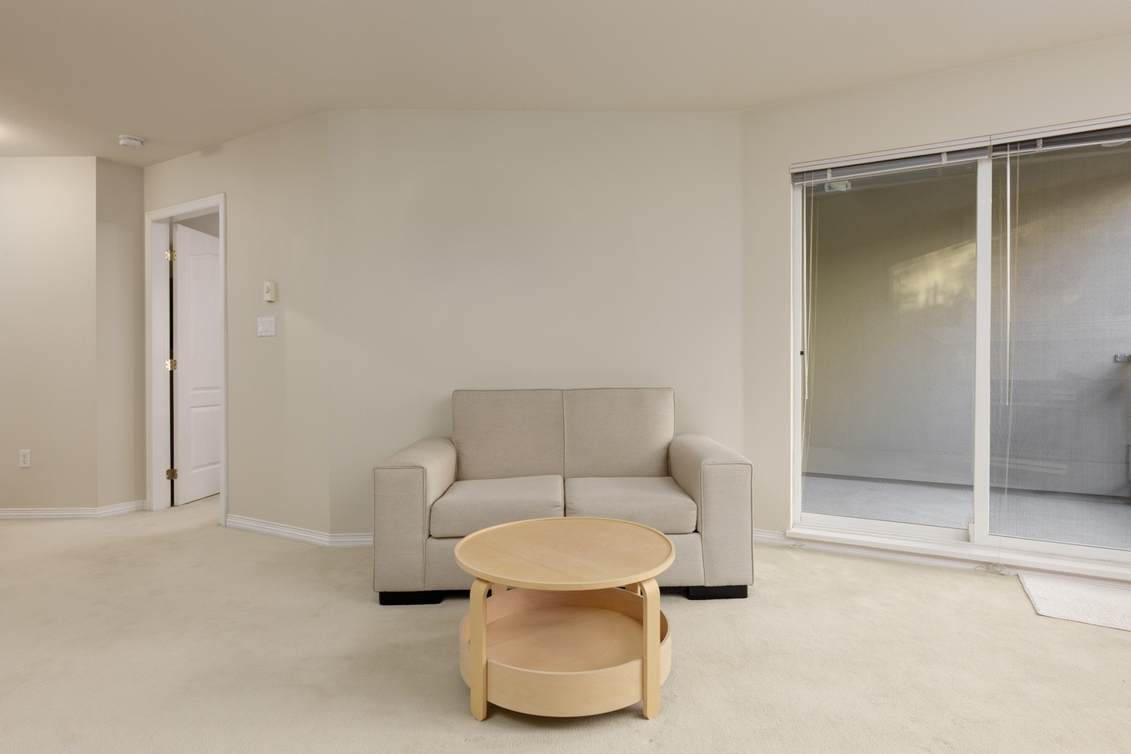 Beige loveseat and round wooden coffee table on a carpeted floor in a 3rd-floor apartment with a sliding glass door.