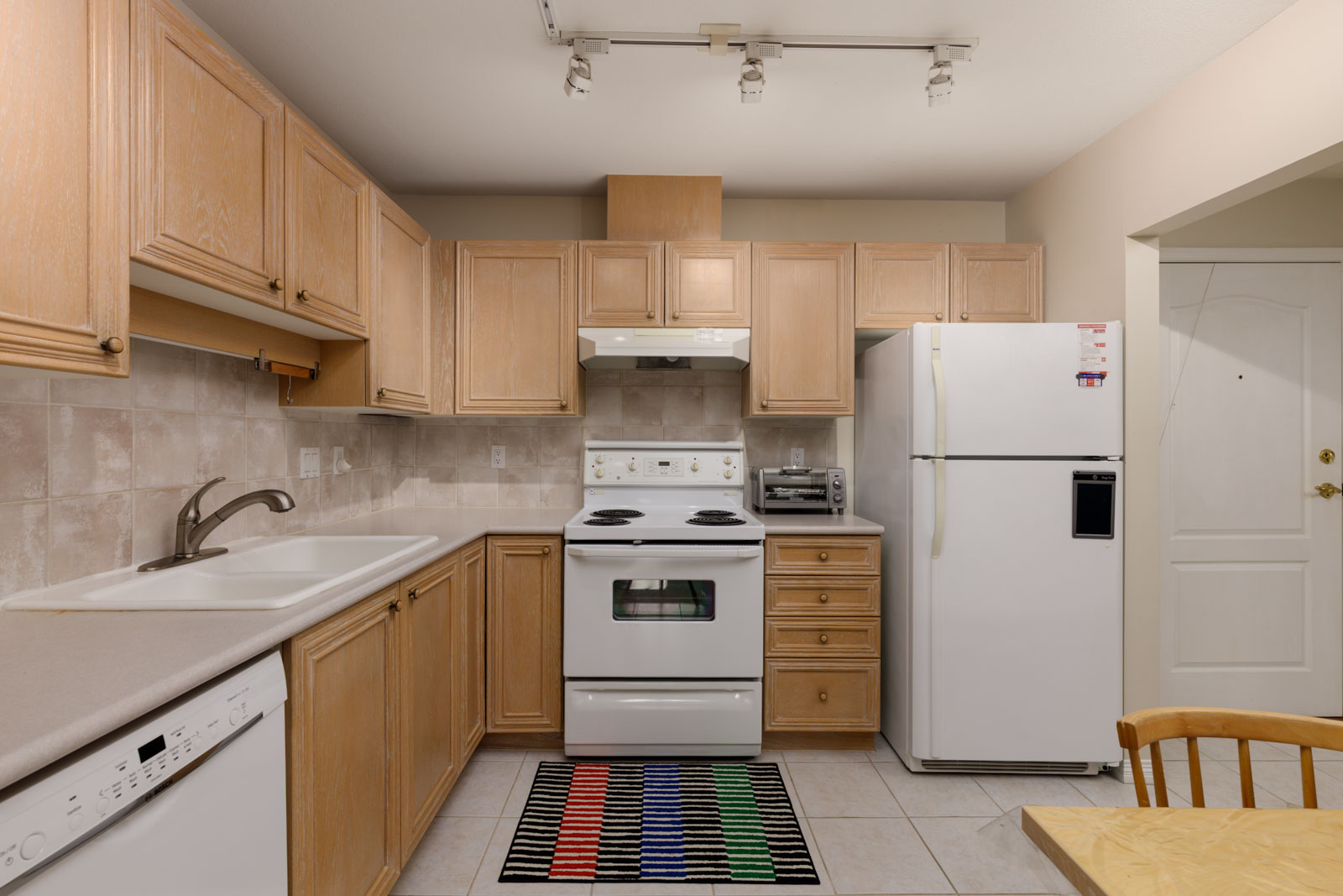 Kitchen at 5683 Hampton Pl with light wood cabinets, white appliances, a tiled backsplash, and a striped floor rug.