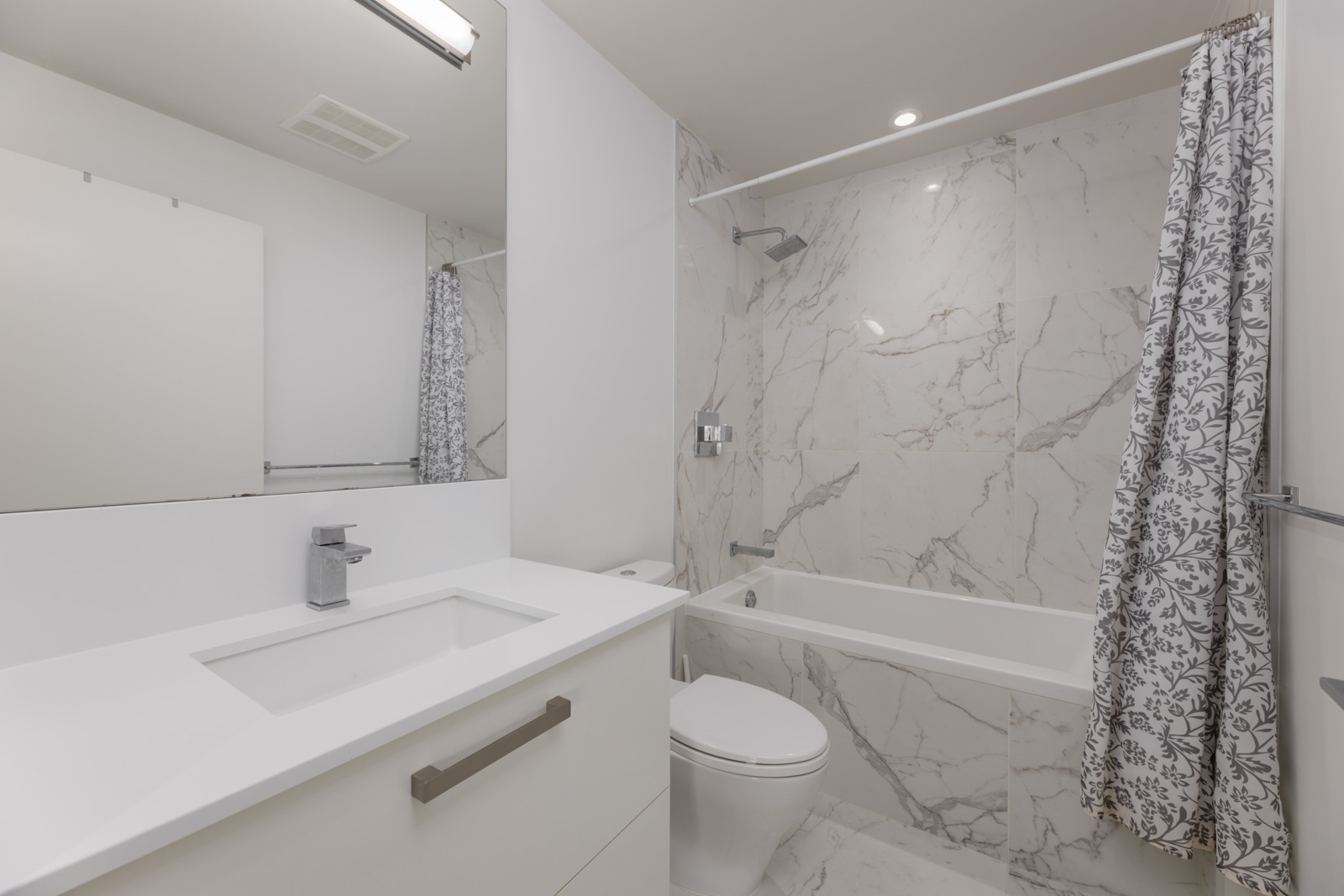 Modern bathroom at 5699 Baillie St with a white vanity, toilet, marble-style tiled bathtub, and floral shower curtain.