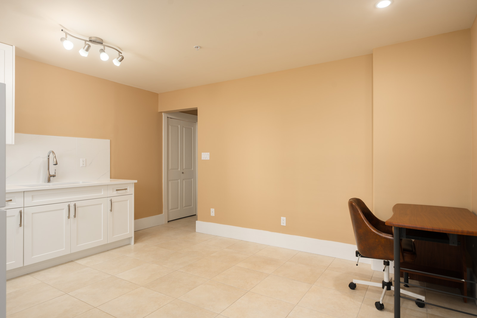 A small kitchen with white cabinets and a sink next to a desk with a brown office chair in a tan-walled West 22nd Ave room.