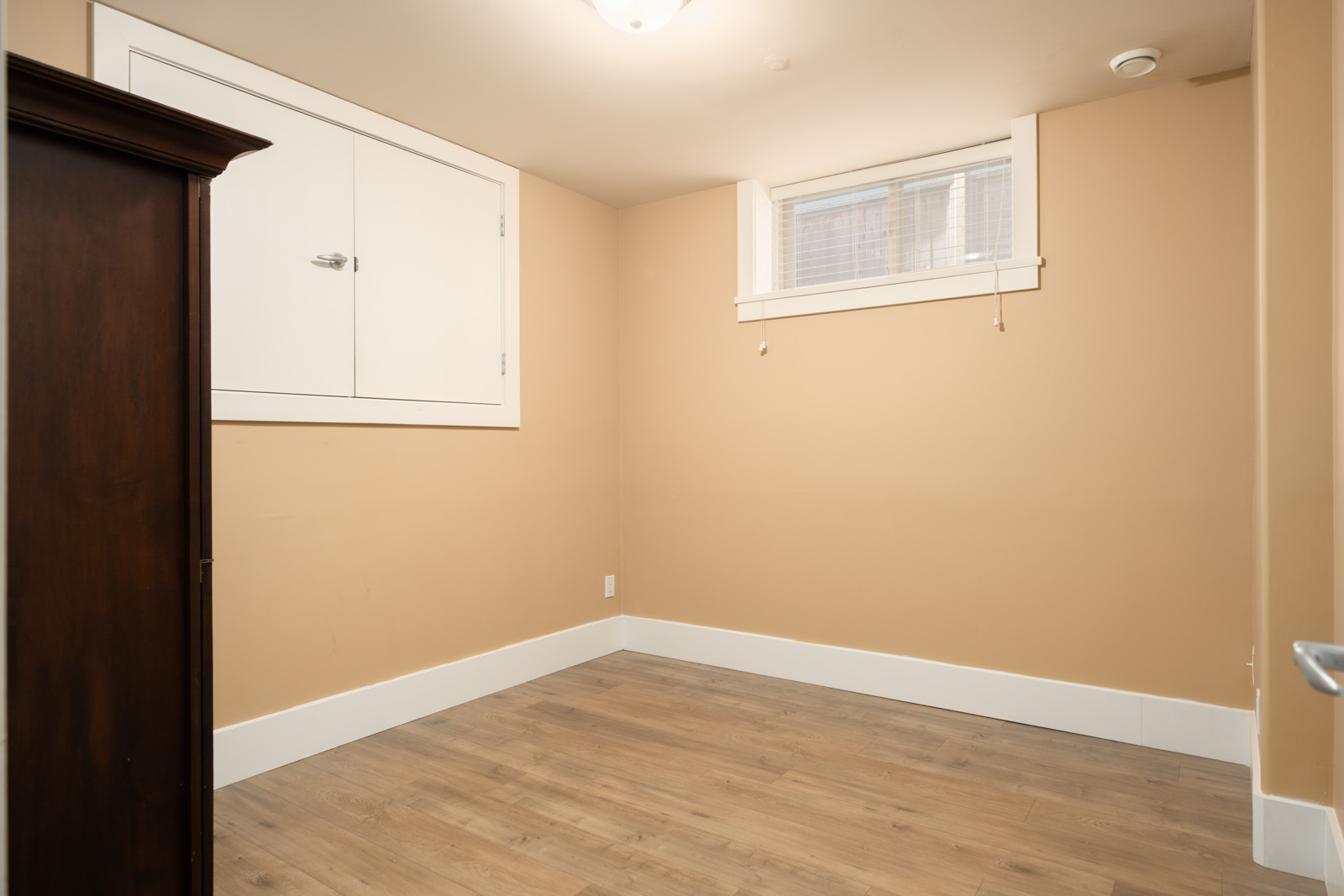 Empty Vancouver basement on West 22nd Ave with beige walls, wood-look flooring, a small window, and a white access door.