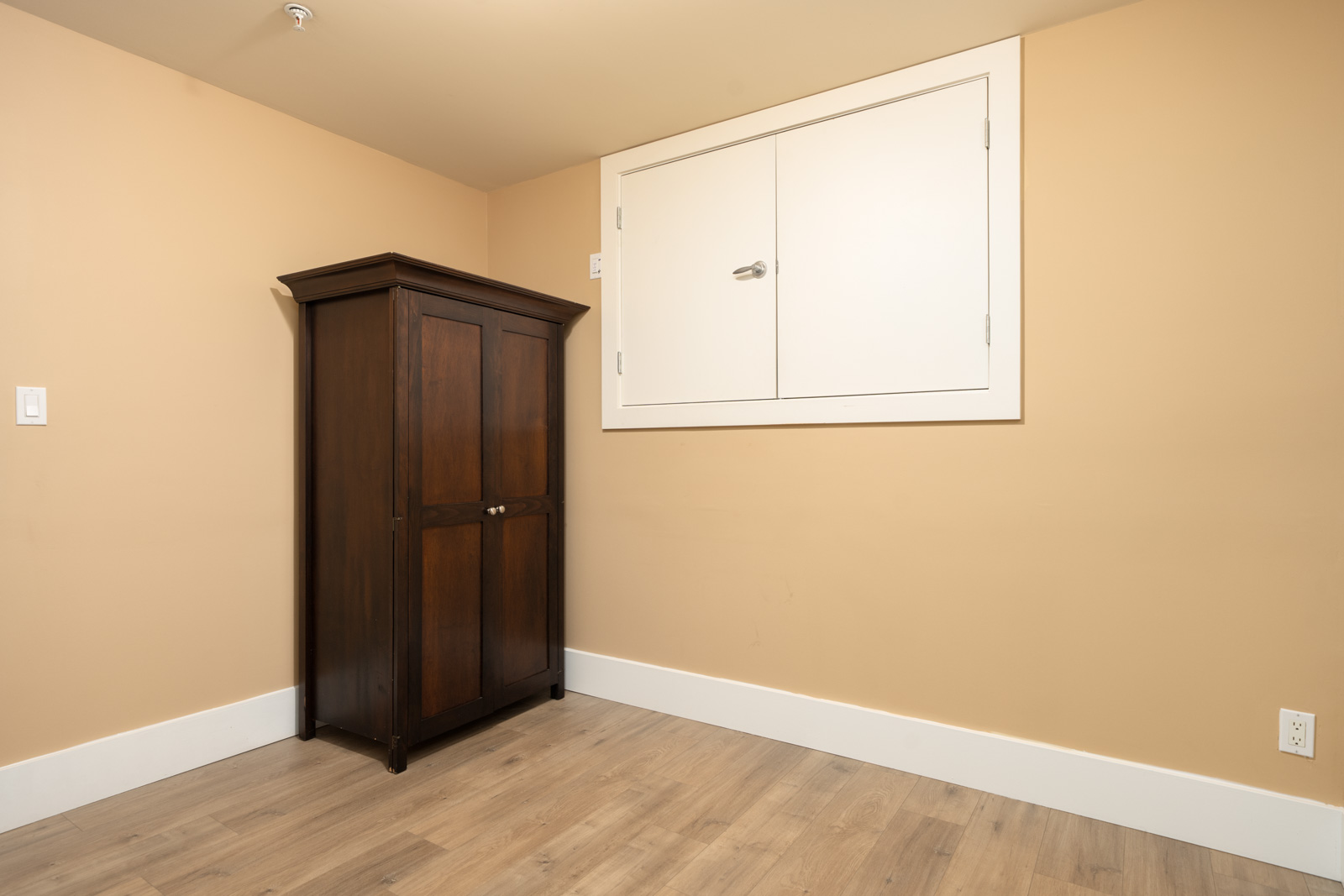 A tall, dark wood cabinet stands against a tan wall next to a white access panel in a Vancouver basement.