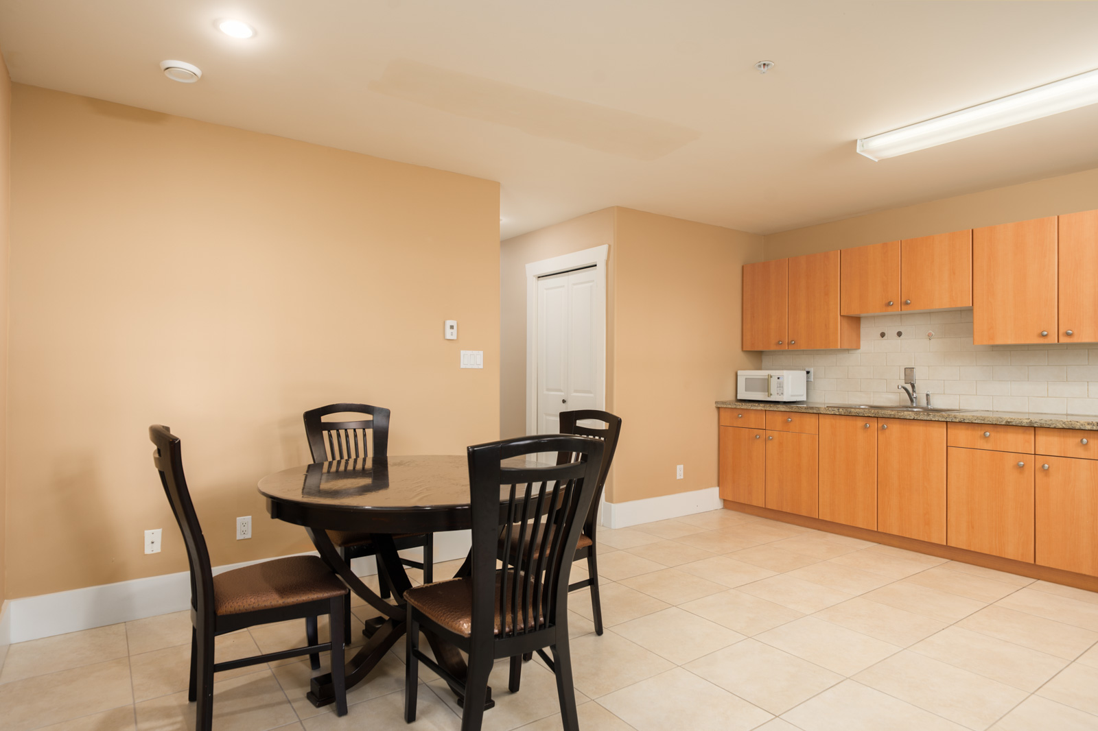 A round black dining table and four chairs in a Vancouver basement kitchen with light wood cabinets and tile floors.