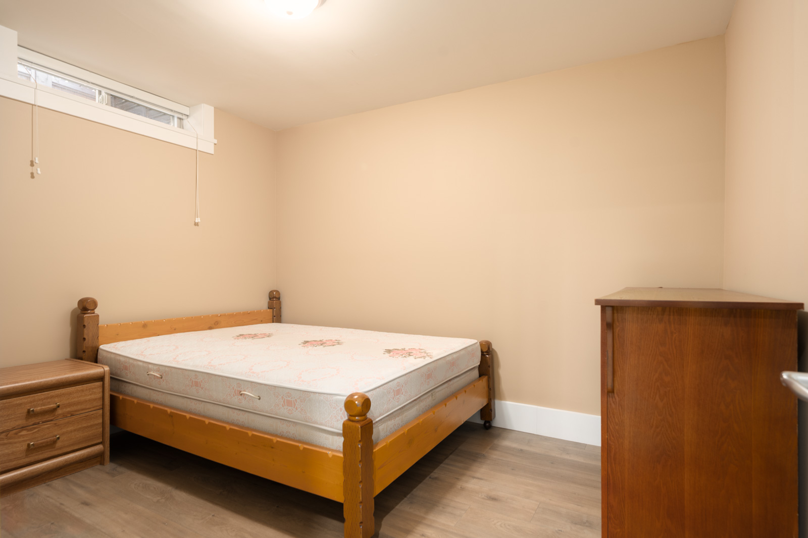 Bedroom with beige walls, wood bed frame, nightstand, and tall dresser in a Yew St, Vancouver basement.